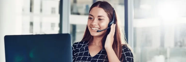 Mulher branca, de cabelos castanhos, atendendo no formato call center e sorrindo na frete da tela do computador.