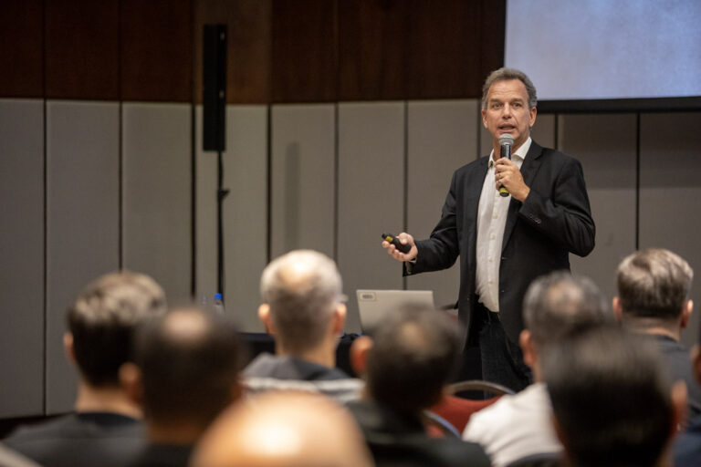 Imagem mostra o professor Alfredo Bravo palestrando em um evento. Ele usa terno preto e camisa branca