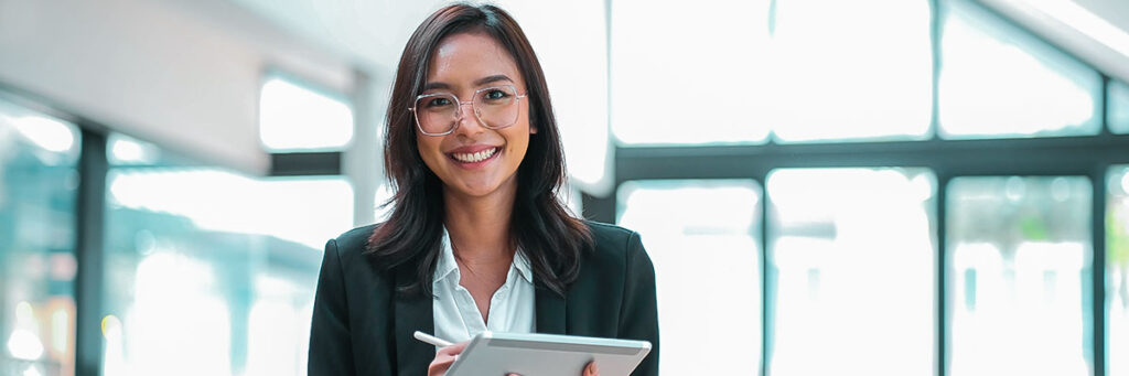 Imagem de mulher branca, de óculos, com roupa social, tablet e sorrindo