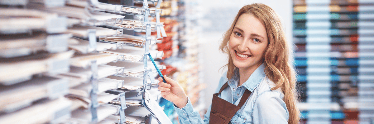 Mulher nova, branca, loira, com avental de atendente contando produtos em um estoque e sorrindo