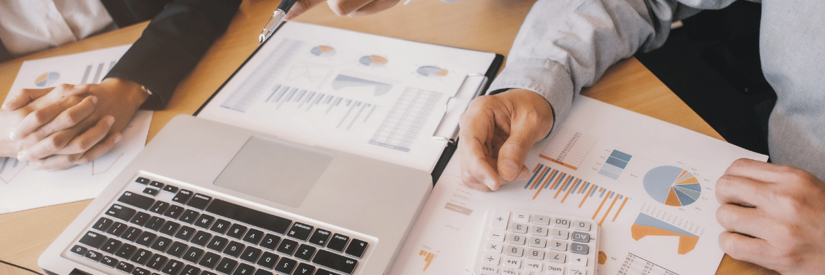 Homens analisando dados em colunas que estão em folhas em cima da mesa, junto com um notebook