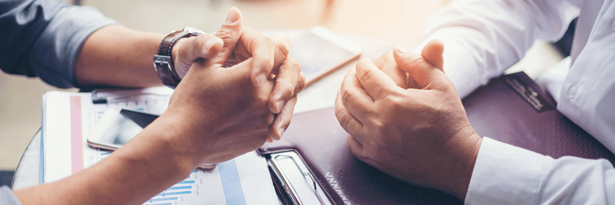 dois homens brancos, vestidos socialmente, frente a frente em uma mesa, negociando