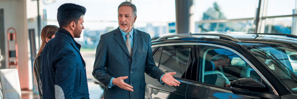 Vendedor, homem mais velho, branco, usando terno e gravata, mostrando um carro para o cliente.