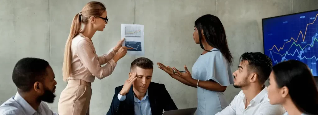 Em reunião de uma empresa, duas mulheres discutindo sobre um gráfico e outros funcionários na mesa olhando. O chefe está escutando tudo de olhos fechado e mão na cabeça
