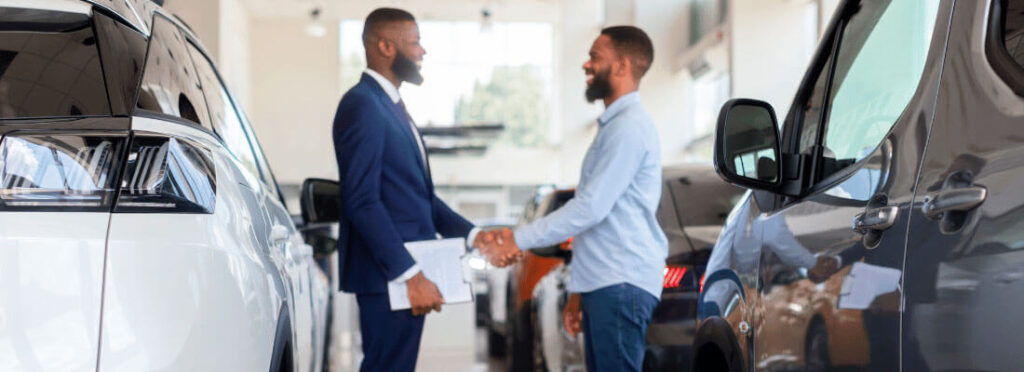 Vendedor, homem negro, de barba, vestindo terno e gravata, apertando a mão de um cliente, em uma concessionária de carros. 