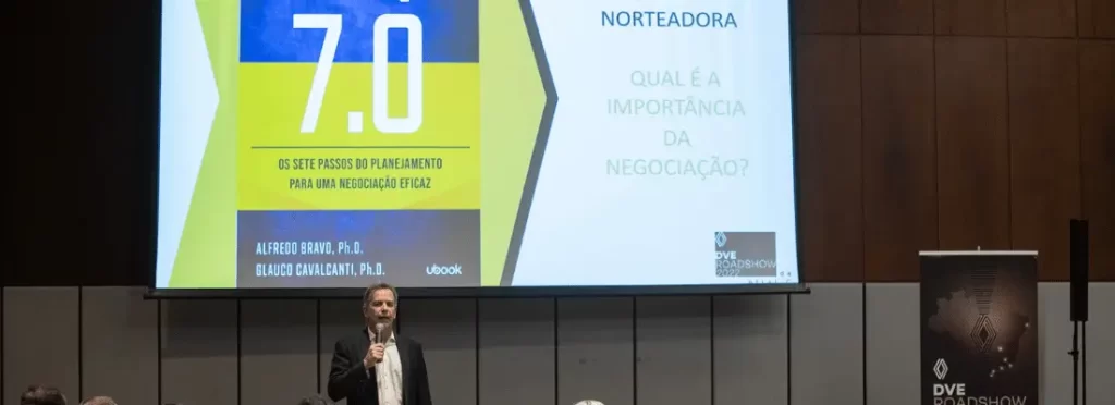 Professor Alfredo Bravo, em pé, falando no microfone em um evento, e atrás um painel com a capa do livro Negociação 7.0.