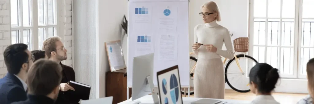 Mulher em pé, vestindo uma roupa social toda branca, falando com uma equipe de pessoas, em uma ambiente corporativo. Ela é branca e tem o cabelo loiro bem claro