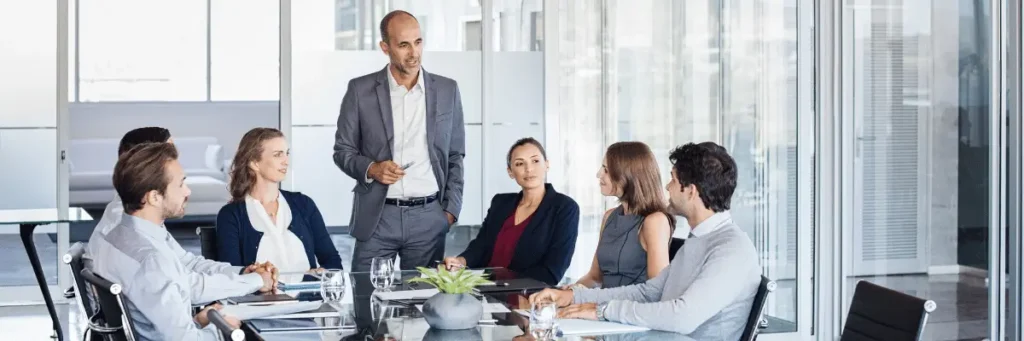 Homem de terno, representando o líder de uma equipe, em pé, falando para algumas pessoas ao redor de uma mesa, em um ambiente corporativo