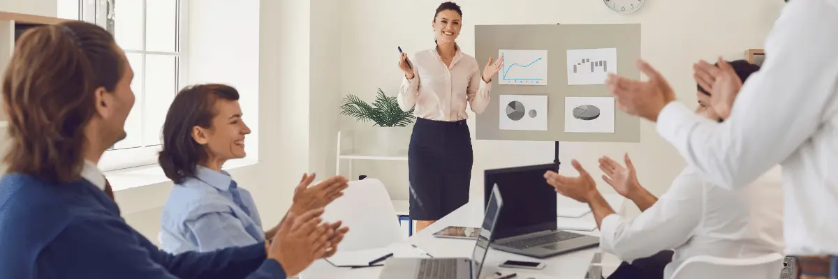 Imagem mostra Mulher em pé, na frente de sua equipe que está sentada ao redor de uma mesa. Eles estão sorrindo, aplaudindo, em um ambiente de escritório.