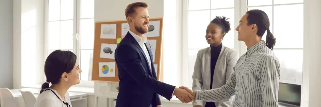 Imagem mostra um homem e uma mulher apertando as mãos, em um ambiente empresarial. Ambos são brancos e vestem roupas sociais.
