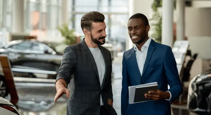 Imagem mostra vendedor e cliente conversando e sorrindo. Ambos usam terno e estão em uma concessionária de automóveis. O vendedor é branco e o cliente é negro