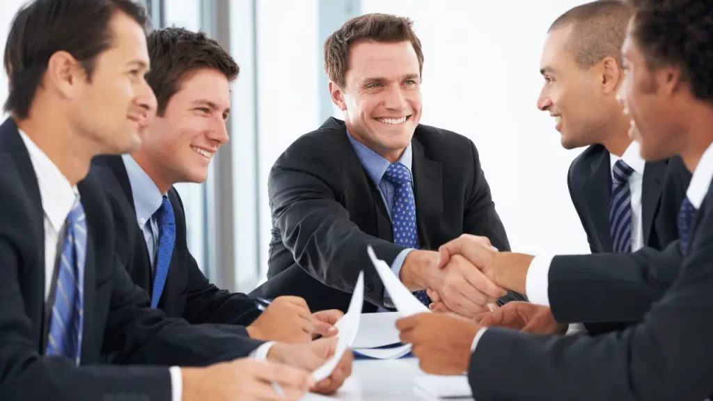 Homens de terno negociando, em uma mesa de um ambiente empresarial. Dois estão sorrindo e apertando as mãos.