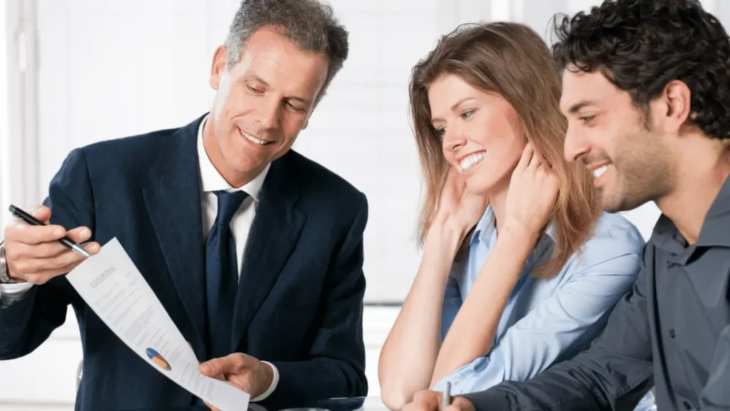 Imagem mostra homem de terno mostrando um documento para um casal de clientes. Todos são brancos e estão sorrindo.