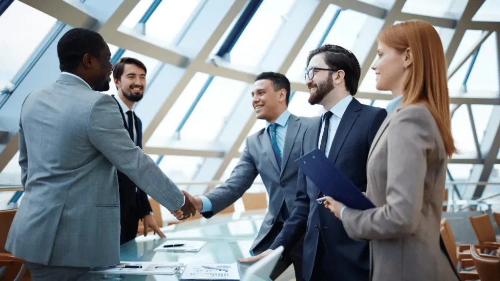 Imagem mostra grupo de empresários em pé, em volta de uma mesa e dois deles estão apertando as mãos. São quatro homens, 3 brancos e um negro. Também há uma mulher branca. Todos estão vestindo roupas sociais.