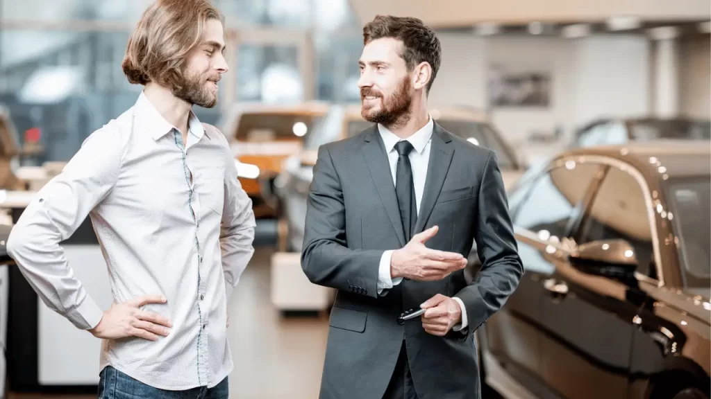 Vendedor falando com o cliente. Ele parece estar explicando algumas coisas sobre um carro que está mostrando. O vendedor está de terno.