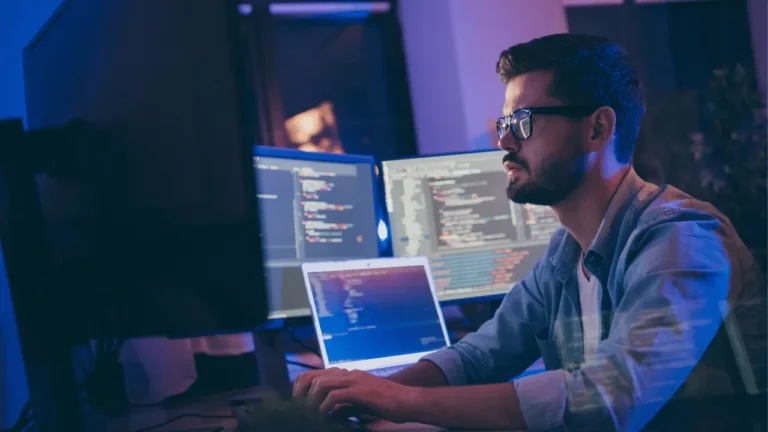 Imagem de homem trabalhando no computador, em uma sala escura, e outros computadores em volta. Ele é branco, usa óculos e veste camisa social.