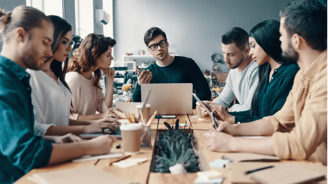 Homem falando com uma equipe, em um ambiente empresarial. Ele está na ponta de uma mesa grande, usa óculos e é branco, assim como todos os outros. Há note books abertos na mesa. A equipe é formada por homens e mulheres.