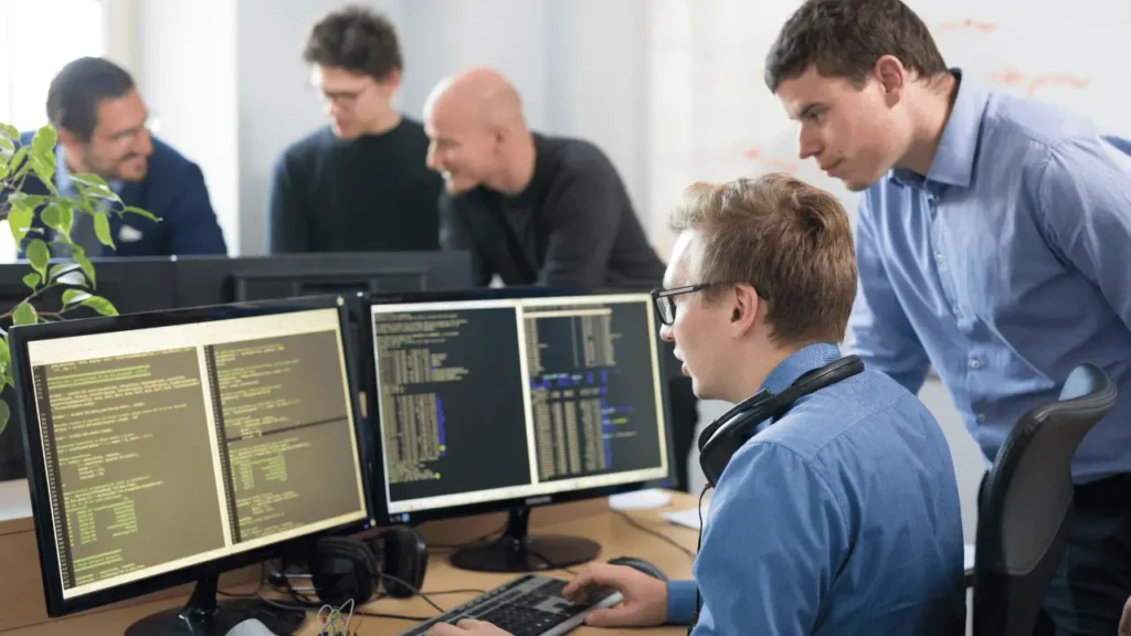 Imagem mostra dois homens analisando duas relas grandes de computador, com uma série de dados. Eles são brancos e estão vestindo camisas  sociais azuis  