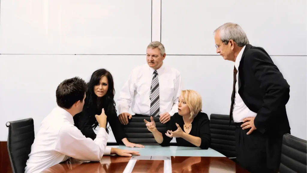 Imagem mostra um grupo de trabalho discutindo em volta de uma mesa. Há três homens e duas mulheres. Todos são brancos e vestem roupas sociais. Dois homens tem cabelos grisalhos e estão em pé.