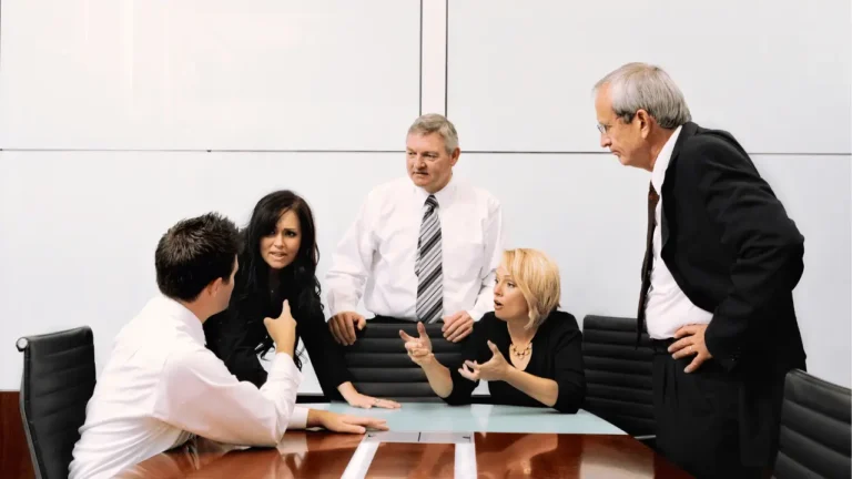 Imagem mostra um grupo de trabalho discutindo em volta de uma mesa. Há três homens e duas mulheres. Todos são brancos e vestem roupas sociais. Dois homens tem cabelos grisalhos e estão em pé.