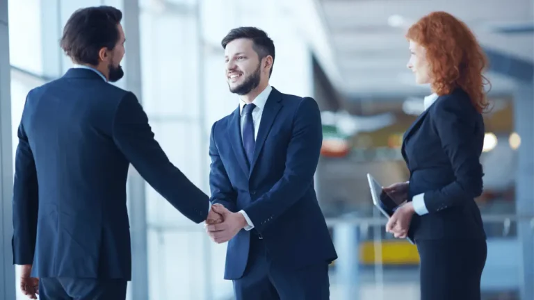 Dois homens apertando as mãos e uma mulher ao lado. Todos são brancos e vestem terno.