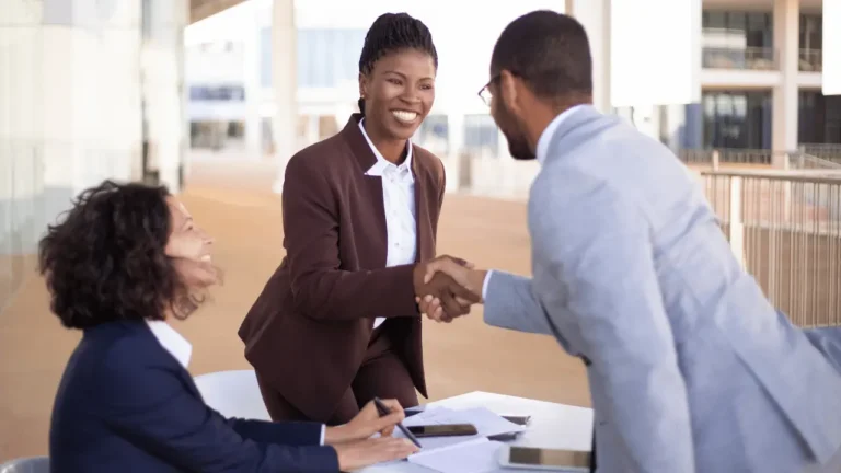 Homem e mulher se cumprimentando em um aperto de mãos. Os dois usam roupas sociais e são negros. Há uma outra mulher ao lado deles, que também veste roupa social e é negra.