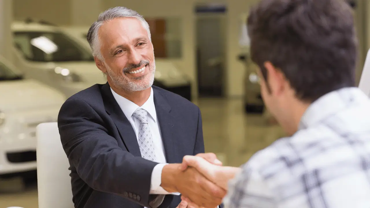 Imagem mostra um vendedor apertando a mão de um cliente. O vendedor aparece de frente. Ele é um homem branco, de barba branca e está usando terno. O cliente aparece de costas e ele é um homem branco e está vestindo uma camisa social branca.