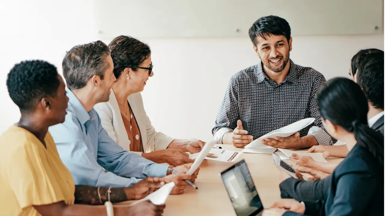 Equipe de trabalho reunida em uma mesa e um homem na ponta da mesa falando com as pessoas. Há homens e mulheres.