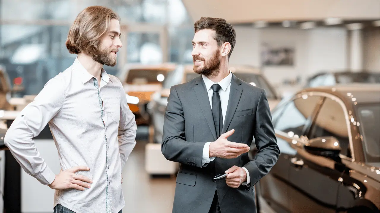 Imagem mostra um vendedor conversando com um cliente, em uma concessionaria de veículos. O vendedor é branco e veste terno. O cliente também é branco e veste uma camisa social de cor clara.