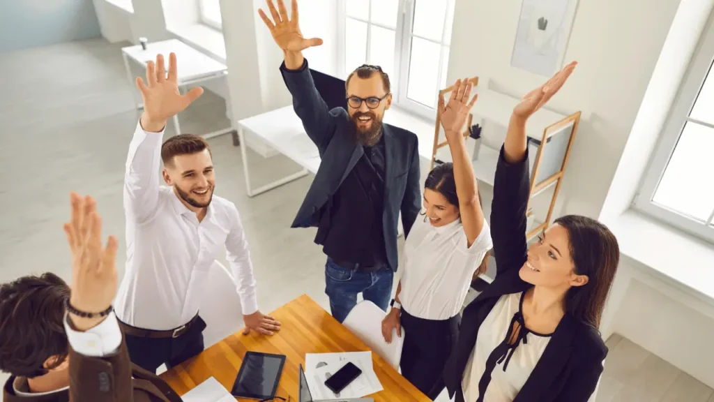 Equipe reunida em torno de uma mesa, que tem um notebook aberto. A equipe é composta por homens e mulheres, todos brancos e estão com os braços levantados, demonstrando empolgação e engajamento.