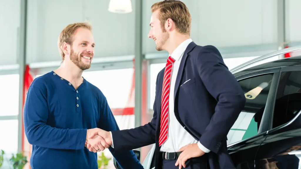 Dois homens apertando as mãos em uma concessionária de automóveis. Os dois homens são brancos, um usa terno e gravata e o outro uma camisa azul.