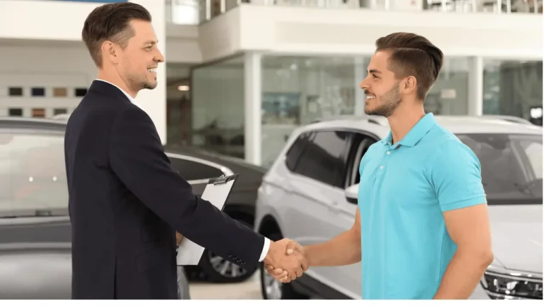 Vendedor e cliente apertando as mãos, em uma concessionária de veículos. O vendedor está de terno escuro e o cliente de camisa polo azul-claro. Ambos são brancos.