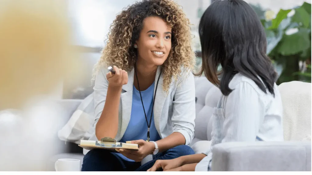 Duas mulheres conversando em um ambiente empresarial.  As duas vestem roupa social. Uma é branca e está de costas na imagem. A outras é negra e está segurando uma prancheta e caneta em outra mão