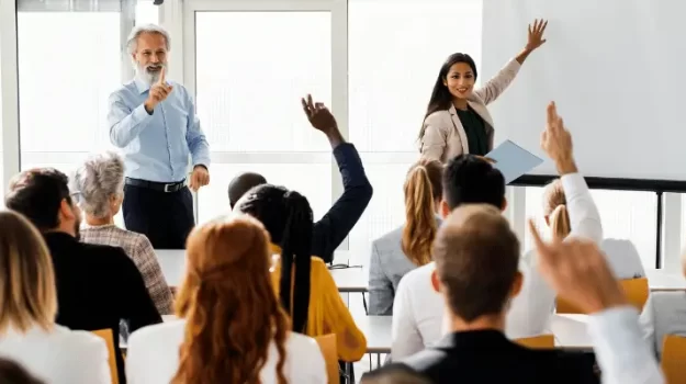 Homem e mulher em pé, na frente de uma turma de pessoas sentadas e com as mãos levantadas.